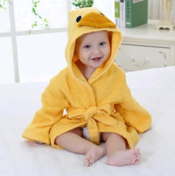 Petit bébé installé assis sur un lit, souriant, et qui porte un peignoir de bain jaune dont la capuche représente une tête de poussin avec deux yeux et un bec orange