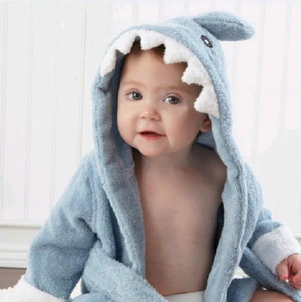 Petit bébé qui porte un peignoir de bain dont la capuche représente une tête de requin, le peignoir est bleu, et autour de la capuche il y a les dents du requin