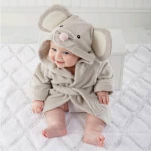 Bébé installé sur un tapis de bain et appuyé sur un petit meuble en bois, porte un peignoir de bain dont la capuche représente une tête de souris , le peignoir est beige , le bébé sourit