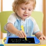 Un petit garçon qui à un stylet jaune dans la main et qui est assis à une table. Il dessine sur une tablette magique à dessin bleu et jaune