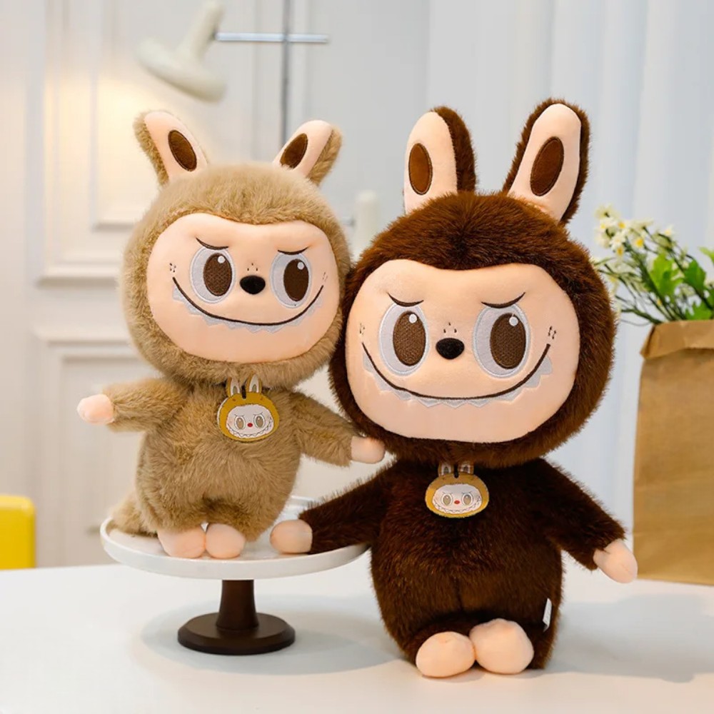 2 peluches style Labubu, une moyenne posée sur un présentoir à gateau de couleur marron clair, et une autre plus grosse marron foncé, posé à coté directement sur la table.