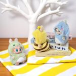 petits costumes en peluche aux formes d’animaux (abeille, dinosaure, baleine) enfilés sur des figurines Labubu, posés sur une surface jaune et blanche.