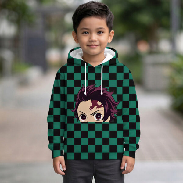 Un petit garçon souriant portant un sweat Demon Slayer Tanjiro à carreaux verts et noirs avec le visage du héros imprimé, posant debout dans une allée extérieure avec des plantes en arrière-plan.