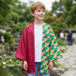 Un enfant porte la veste de kimono de Giyu Tomioka Demon Slayer pour enfant, avec une moitié rouge et une moitié verte à motifs, debout sur un pont en bois dans un jardin avec un étang et des arbres derrière.