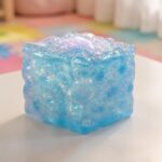 Un jouet anti-stress sensoriel Needoh à paillettes bleu en forme de cube, posé sur une table claire dans un décor doux et coloré.