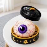 Un jouet garçon anti-stress Halloween en forme d'œil rougi posé dans une boîte noire et jaune, avec un gros œil blanc aux veines rouges et une pupille violette.