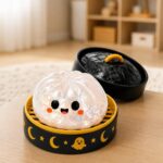 Un jouet garçon anti-stress Halloween petit fantôme joufflu translucide avec visage souriant, posé dans une boîte ronde noire décorée de lunes et de petits fantômes sur une table.