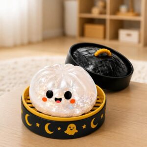 Un jouet garçon anti-stress Halloween petit fantôme joufflu translucide avec visage souriant, posé dans une boîte ronde noire décorée de lunes et de petits fantômes sur une table.