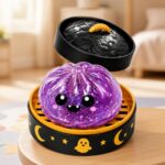 Un jouet garçon anti-stress Halloween violet aux joues roses en forme de fantôme violet, posé dans une boîte noire et orange décorée de lunes et de fantômes.