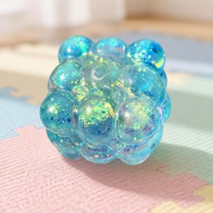 Un jouet squishy garçon en cube rempli de billes brillantes bleu transparent, posé sur un tapis coloré en mousse.