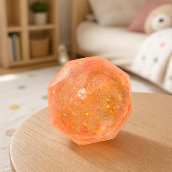 Un Jouet Squishy Needoh garçon anti-stress en balle à facettes orange translucide, posé sur une petite table en bois dans un salon doux et lumineux.