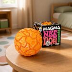 Un jouet Squishy Needoh garçon anti-stress en balle texture lave orange posé sur une table, avec sa boîte noire et rose en arrière-plan dans une chambre lumineuse.