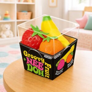 Un jouet Squishy Needoh garçon anti-stress en fraise et banane dans une boîte transparente sur une petite table, avec une fraise rouge, une banane jaune et une orange.
