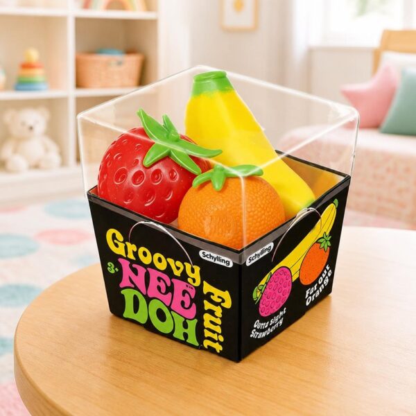 Un jouet Squishy Needoh garçon anti-stress en fraise et banane dans une boîte transparente sur une petite table, avec une fraise rouge, une banane jaune et une orange.