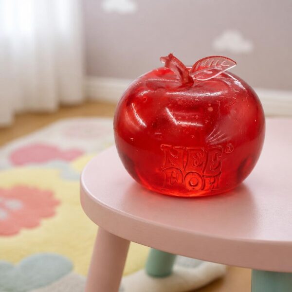 Un jouet squishy Needoh garçon anti-stress en pomme pailletée rouge, posé sur une petite table rose dans une chambre douce et lumineuse.