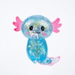 Un jouet squishy axolotl maltose bleu transparent, avec des paillettes colorées, de grands yeux noirs, des branchies roses et une queue, présenté sur un fond blanc.